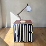 Load image into Gallery viewer, A modern table lamp with a white conical shade and a dark metal articulated arm sits on top of a cube-shaped side table. The table features the word 'GAULTIER' in large black letters on a cream background.
