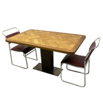 Load image into Gallery viewer, A mid-century modern dining table set featuring a rectangular wooden table with a herringbone pattern top and a black pedestal base, accompanied by two chairs with chrome tubular frames and burgundy upholstered seats, shown on a white background.
