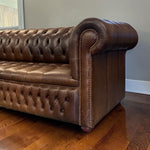 Load image into Gallery viewer, A close-up of a brown leather Chesterfield sofa with deep button tufting, rolled arms, and nailhead trim, shown on a hardwood floor against a gray wall.
