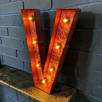 Load image into Gallery viewer, A large decorative letter V marquee light with a warm orange crackled glass face and glowing bulbs, leaning on a wooden surface against a gray brick wall.
