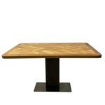 Load image into Gallery viewer, A rectangular dining table with a light brown and beige herringbone wood top and a black metal pedestal base, shown against a plain white background.
