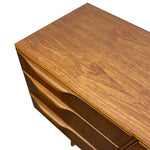 Load image into Gallery viewer, Close-up of a mid-century modern wooden dresser or desk with a warm walnut finish, showing the top surface, drawer front, and curved drawer pulls against a white background.

