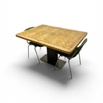 Load image into Gallery viewer, A rectangular wooden dining table with a herringbone-patterned wood top and a dark pedestal base, accompanied by two dark green chairs with metal legs, shown on a white background.
