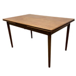 Load image into Gallery viewer, A mid-century modern wooden dining table with a warm brown finish, featuring tapered legs and a visible center pull drawer, shown against a plain white background.
