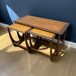 Load image into Gallery viewer, A set of three nested wooden nesting tables with a mid-century modern design, featuring curved legs and a warm medium-brown wood finish, set against a dark blue wall and light wood floor.
