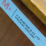 Load image into Gallery viewer, Close-up of a light blue printed label or sticker placed on a wooden surface, showing partial red lettering and the text 'A.H.

