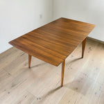Load image into Gallery viewer, A mid-century modern wooden dining table with a rich brown grain finish and tapered legs, shown on a light wood plank floor against a white wall.

