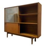 Load image into Gallery viewer, Mid-century modern wooden bookcase with a warm brown wood finish, featuring a glass-front cabinet on the left, two open shelves, and a lower cabinet door, shown on a plain white background.
