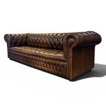 Load image into Gallery viewer, A classic brown leather Chesterfield sofa with deep button tufting, rolled arms, and nailhead trim, shown from a three-quarters angle on a white background.
