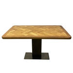 Load image into Gallery viewer, A rectangular dining table with a light brown wood herringbone-patterned top and a black metal pedestal base, shown against a plain white background.
