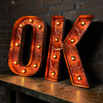 Load image into Gallery viewer, Large decorative letters spelling "OK" in a glowing marquee style, with an orange cracked-metal look and built-in light bulbs, displayed against a dark brick wall on a wooden surface.
