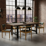 Load image into Gallery viewer, A modern dining table set in a loft-style room with brick walls, large industrial windows, and three black pendant lights. The set includes a rectangular wooden table with a black metal base and four matching wooden chairs with black upholstered seats.
