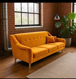 Load image into Gallery viewer, A mustard yellow velvet loveseat sofa with tufted backrest and two matching throw pillows, placed in a loft-style room with brick walls, wood floors, a window, a floor lamp, and a potted plant.
