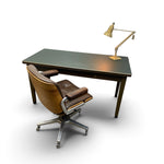 Load image into Gallery viewer, A mid-century modern writing desk with a dark green top and wood legs, paired with a brown leather swivel chair and a brass adjustable desk lamp, shown on a white background.
