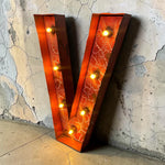 Load image into Gallery viewer, A large decorative letter V marquee light with warm glowing bulbs, featuring a cracked orange-red metallic finish and dark brown edging, leaning against a textured concrete wall.
