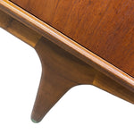 Load image into Gallery viewer, Close-up of a mid-century modern wooden cabinet or sideboard, showing the warm brown wood grain of the drawer front, trim, and tapered leg against a white background.
