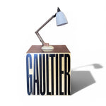 Load image into Gallery viewer, A modern table lamp with a light blue conical shade and a dark metal articulated arm, mounted on a cube-shaped wooden side table. The table features the brand name 'GAULTIER' in large black and blue block letters on the front, set against a clean white background with a soft shadow.
