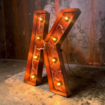 Load image into Gallery viewer, A large decorative letter K floor lamp with a glowing orange marbled pattern and warm light bulbs, set against a rustic, distressed concrete and metal wall.
