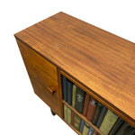 Load image into Gallery viewer, A close-up of a mid-century modern wooden sideboard or cabinet, showing a warm walnut-toned wood grain finish, a single cabinet door with a small round pull, and open shelving holding books, set against a plain white background.
