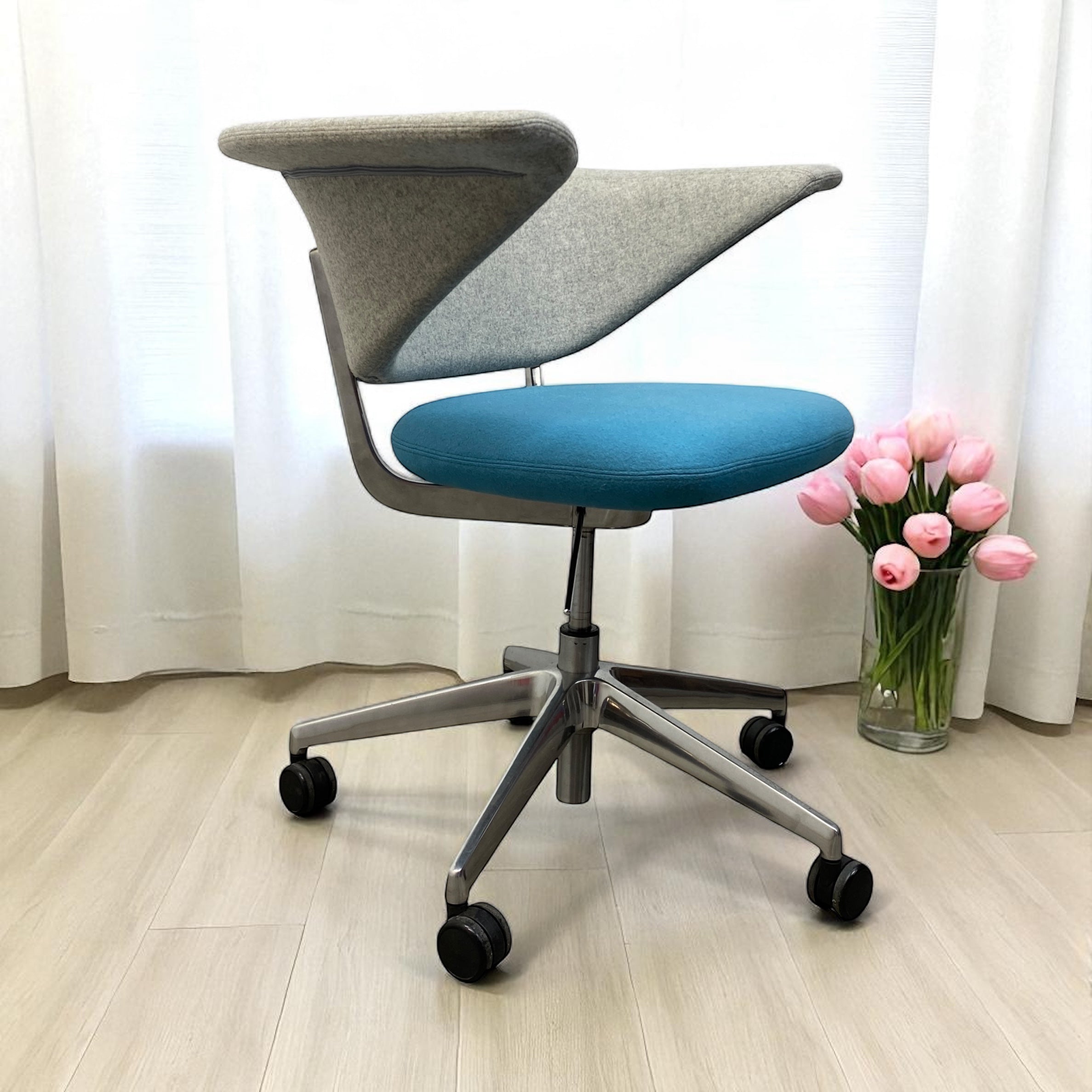 Modern office chair with blue seat and gray backrest on a light wooden floor, next to a vase of pink flowers.