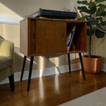 Load image into Gallery viewer, Mid-century modern wooden sideboard cabinet with black tapered legs and gold-tone tips, shown in a styled room setting with a record player on top, books in the open shelf, a potted plant, and part of an armchair.
