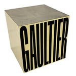 Load image into Gallery viewer, A cube-shaped metallic box with a brushed silver top and sides, and a cream-colored front face featuring the word 'GAULTIER' in large black letters.
