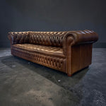 Load image into Gallery viewer, A classic brown leather Chesterfield sofa with deep button tufting and rolled arms, photographed in a dark studio setting with a concrete floor.
