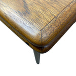 Load image into Gallery viewer, Close-up of a mid-century modern wooden table corner with a polished medium-brown wood grain finish and a dark metal spike leg on a white background.
