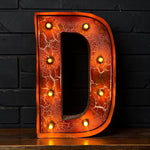 Load image into Gallery viewer, A large decorative letter D marquee light with a cracked orange and red metallic finish, illuminated with warm yellow bulbs, standing on a wooden surface against a dark brick wall.
