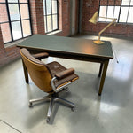 Load image into Gallery viewer, A mid-century modern desk setup featuring a dark green writing desk with a brass desk lamp and a brown leather swivel chair with a chrome five-star base, set in an industrial loft room with brick walls and concrete floors.

