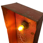 Load image into Gallery viewer, A close-up of a rustic industrial-style light fixture with a glowing warm yellow bulb set inside a reddish-brown metal box with a textured orange-red crackled glass panel.
