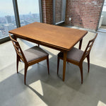 Load image into Gallery viewer, A mid-century modern wooden dining table with two matching wooden chairs featuring gray upholstered seats, set in a bright room with a concrete floor and large windows overlooking a city skyline.
