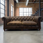 Load image into Gallery viewer, A classic brown leather Chesterfield sofa with deep button tufting and rolled arms, positioned in a spacious industrial loft with exposed brick walls and large windows.
