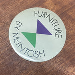 Load image into Gallery viewer, A round white sticker or decal with the text 'FURNITURE BY McIntosh' in black around a central green and purple triangle, placed on a brown textured surface.
