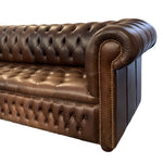 Load image into Gallery viewer, A close-up of a classic Chesterfield sofa in dark brown tufted leather, showing the rolled arm with nailhead trim, button-tufted backrest, and seat cushion against a plain white background.

