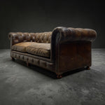 Load image into Gallery viewer, A vintage Chesterfield-style leather sofa in distressed dark brown, featuring deep button tufting, rolled arms, and dark wooden feet, photographed in a studio setting on a dark gray floor against a dark gray background.
