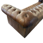 Load image into Gallery viewer, A close-up of a brown tufted leather sofa corner, showing the button-tufted armrests and back cushion on a plain white background.
