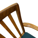 Load image into Gallery viewer, Close-up of a wooden chair back with a medium brown walnut finish, featuring a curved top rail, vertical spindles, and part of a dark teal seat cushion against a white background.

