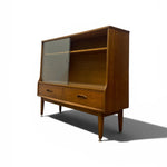 Load image into Gallery viewer, Mid-century modern wooden hutch cabinet with a glass-front upper cabinet, open shelf, two drawers, and tapered legs, shown on a white background with a shadow.
