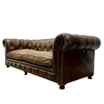 Load image into Gallery viewer, A vintage Chesterfield loveseat sofa upholstered in distressed brown leather, featuring deep button tufting, rolled arms, nailhead trim, and dark wooden feet, shown against a plain white background.

