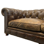 Load image into Gallery viewer, A close-up of a classic Chesterfield sofa in distressed brown leather, showing the tufted backrest, nailhead trim, and a thick seat cushion against a plain white background.
