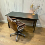 Load image into Gallery viewer, A mid-century modern brown leather swivel office chair with a matching footrest, positioned next to a dark wood desk with a brass desk lamp, set against a light wood-paneled wall and wooden floor.
