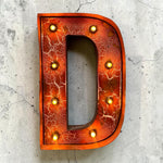 Load image into Gallery viewer, A decorative marquee-style letter D sign with an orange and red cracked metallic finish and warm glowing light bulbs around the border, photographed against a light concrete surface.
