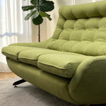 Load image into Gallery viewer, A modern chaise lounge sofa upholstered in textured olive green fabric, shown in a bright living room setting with a potted fiddle-leaf fig plant and sheer curtains in the background.
