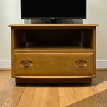 Load image into Gallery viewer, A mid-century modern wooden TV stand with a warm honey finish, featuring a single drawer with oval handles and an open shelf, shown holding a television in a room with light-colored walls and a wood floor.
