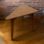 Load image into Gallery viewer, A triangular wooden table with tapered legs, shown in a room setting with a brick wall and hardwood floor.

