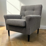 Load image into Gallery viewer, A modern gray upholstered armchair with a tufted backrest and black legs, shown in a room with a light wood herringbone floor and white wall.
