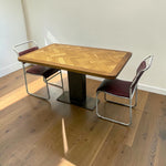 Load image into Gallery viewer, A vintage-style wooden dining table with a herringbone-patterned wood top and a black pedestal base, accompanied by two chrome-framed chairs with burgundy upholstered seats, set on a wooden floor against a plain wall.
