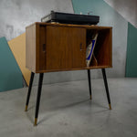 Load image into Gallery viewer, Mid-century modern wooden cabinet with black tapered legs and brass feet, holding a turntable and records, set against a geometric teal and gray wall.
