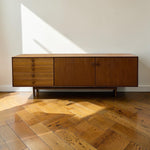Load image into Gallery viewer, Mid-century modern wooden sideboard cabinet with three drawers on the left and two doors on the right, set against a white wall on a herringbone wood floor with sunlight streaming in.
